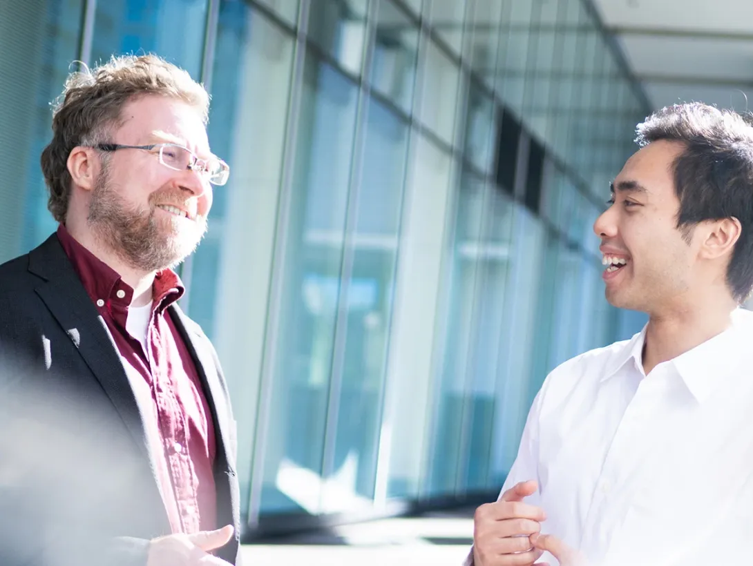 Two smiling male students talk in front of a building