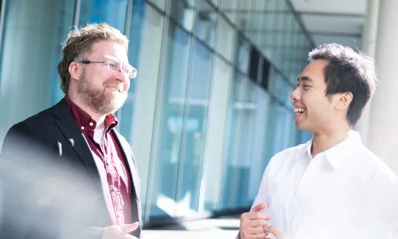 Two smiling male students talk in front of a building