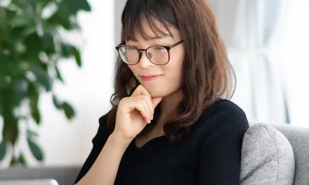A woman with glasses sitting on a sofa, thoughtfully looking at a tablet while resting her chin on her hand.