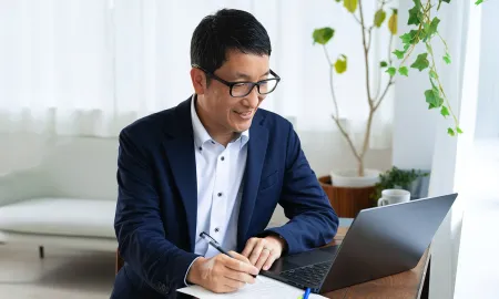 A business school student smiling and taking notes while looking at his laptop screen.
