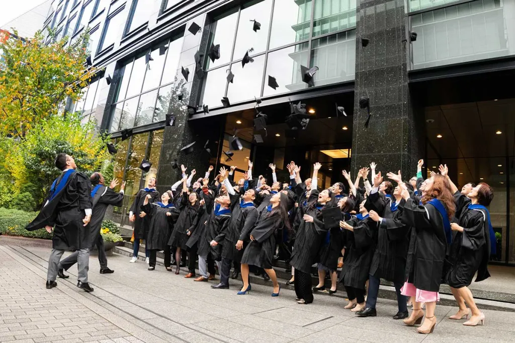 a group of freshly graduated MBA students throw their graduation caps up in the air just outside a building