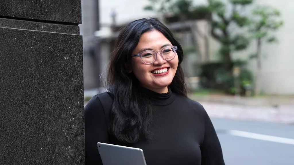 A smiling woman with glasses and long dark hair, wearing a black turtleneck and holding a tablet outdoors.