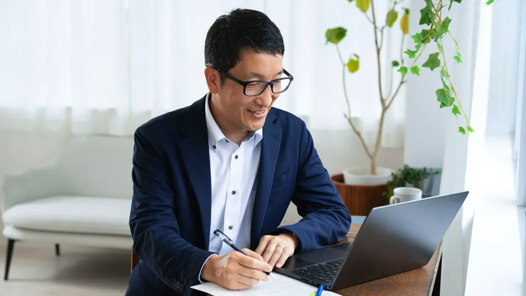 A business school student smiling and taking notes while looking at his laptop screen.