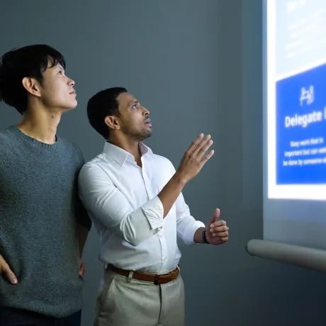 Two MBA students stand in a modern learning space, focusing a digital presentation projected onto a wall. One student gestures toward a slide, explaining a management framework to his peer.