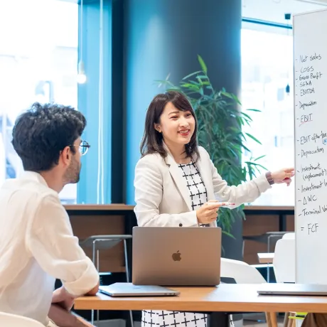 A smiling student in a white blazer points to a whiteboard listing financial terms like EBITDA and FCF while a student at the same table listens.